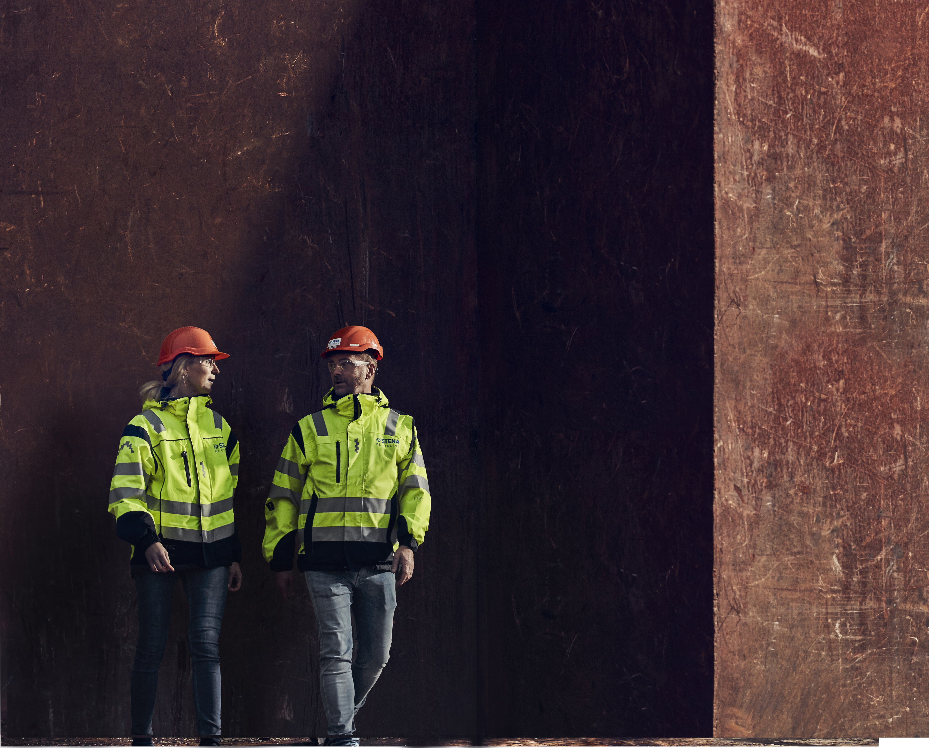 Two Stena Metall Group employees - one male and one female - talk together wearing hi-vis protective clothing.