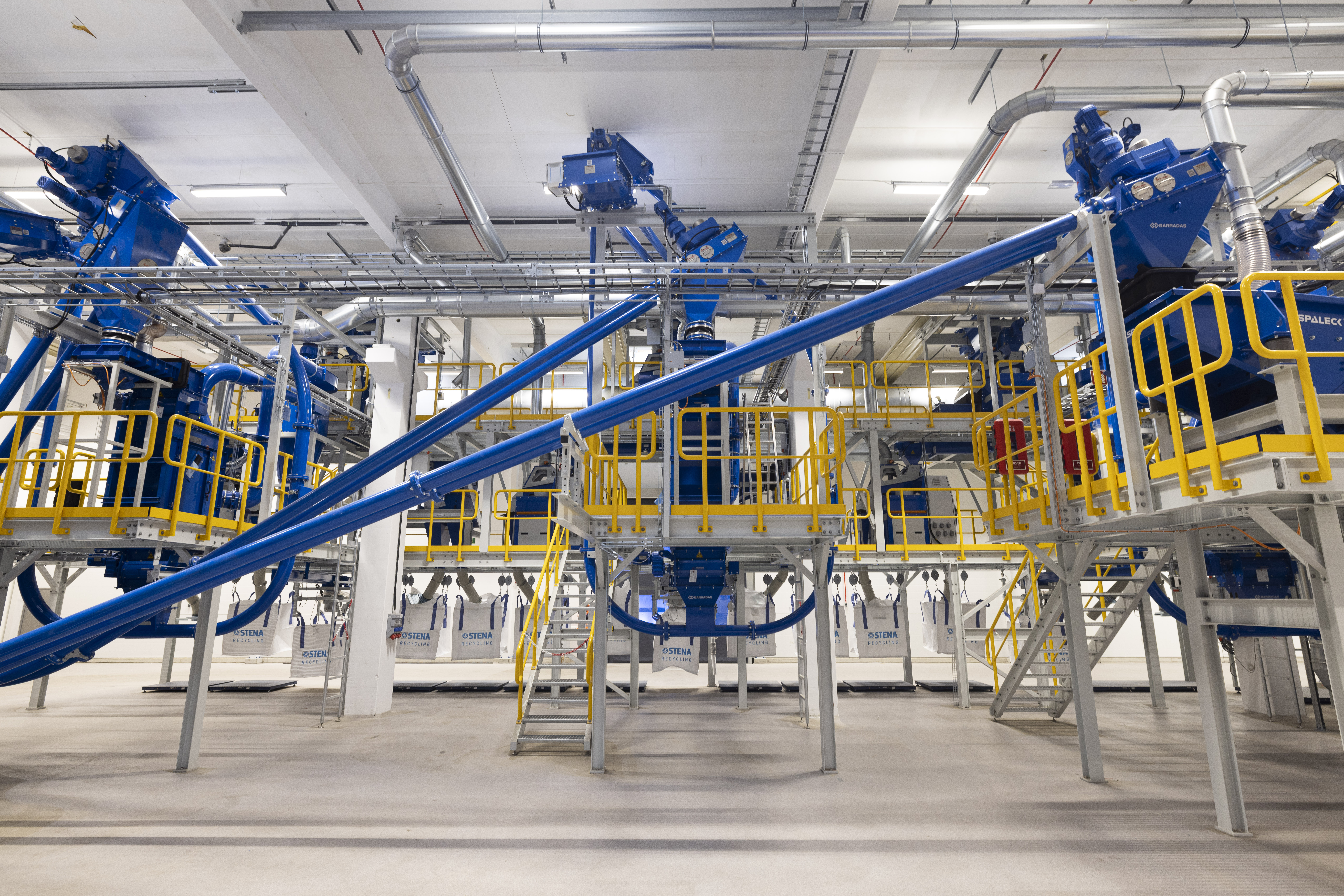 The interior of a battery recycling facility, with yellow, blue and white machinery, pipes and landings.