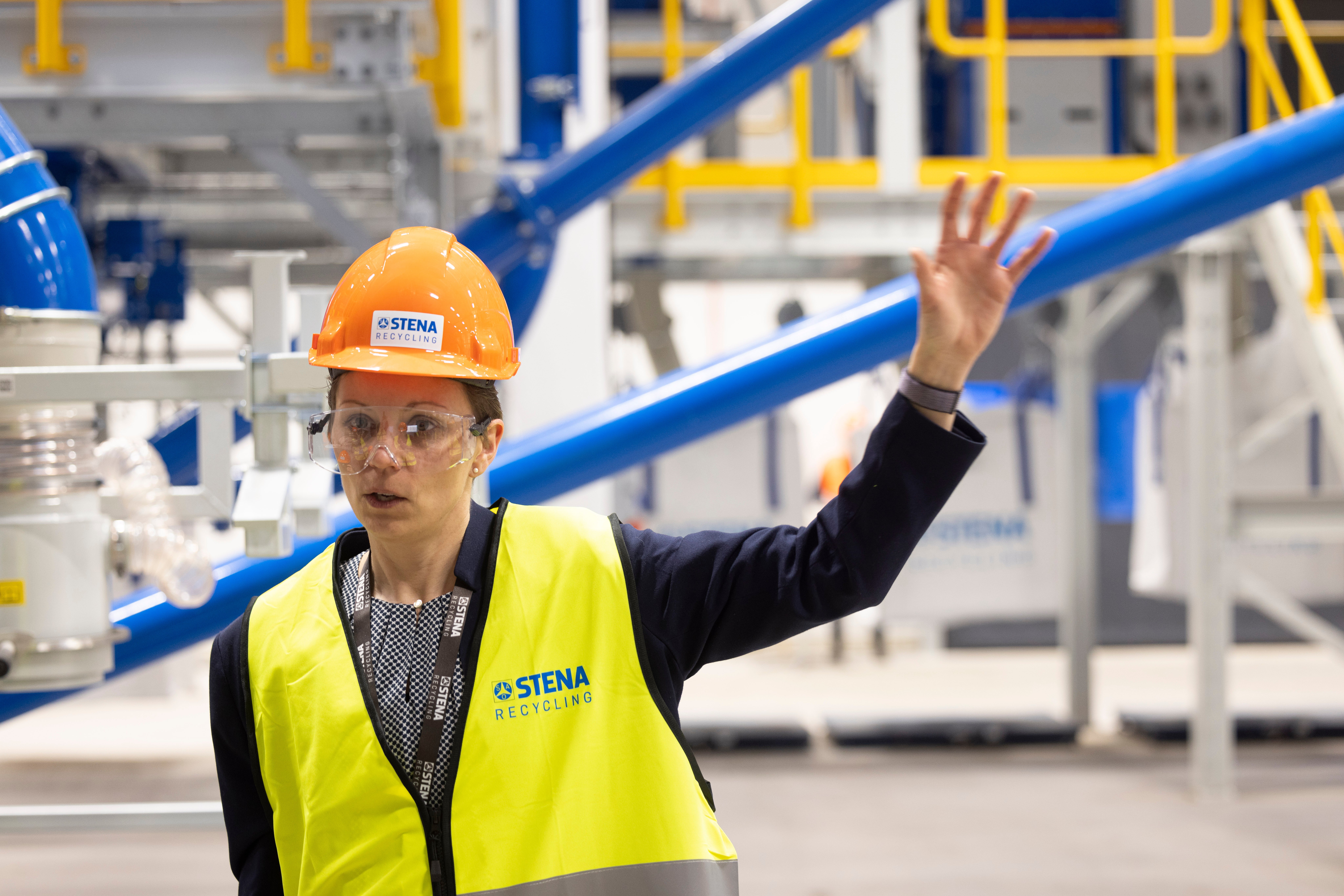 A female Stena Recycling employee in hi-vis protective gear, raising her left hand. In the background, yellow, blue and white machinery is visible.