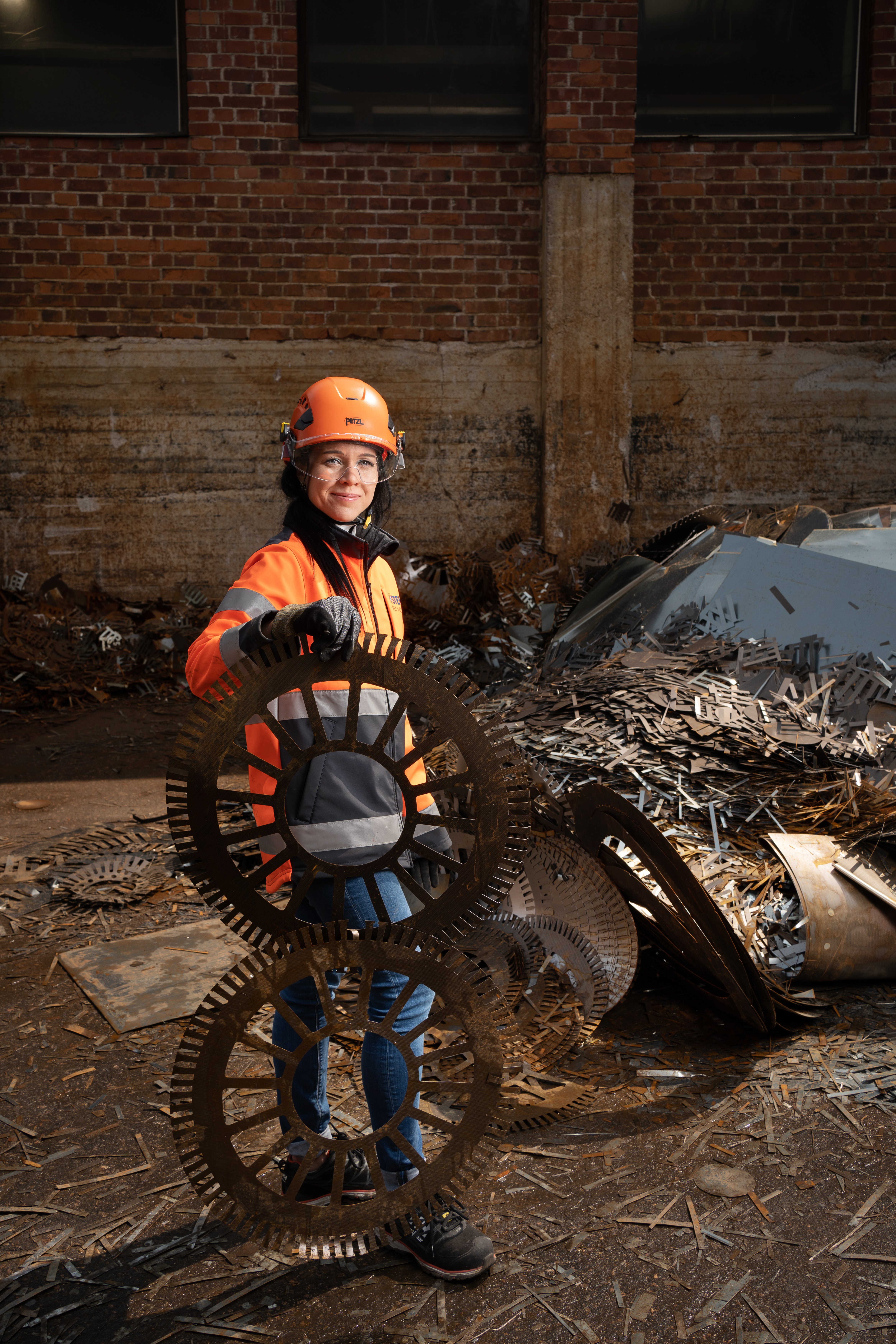 A female Stena Recycling employee in hi-vis protective gear, standing in front of a pile with metal scrap, holding round pieces of metal scrap.