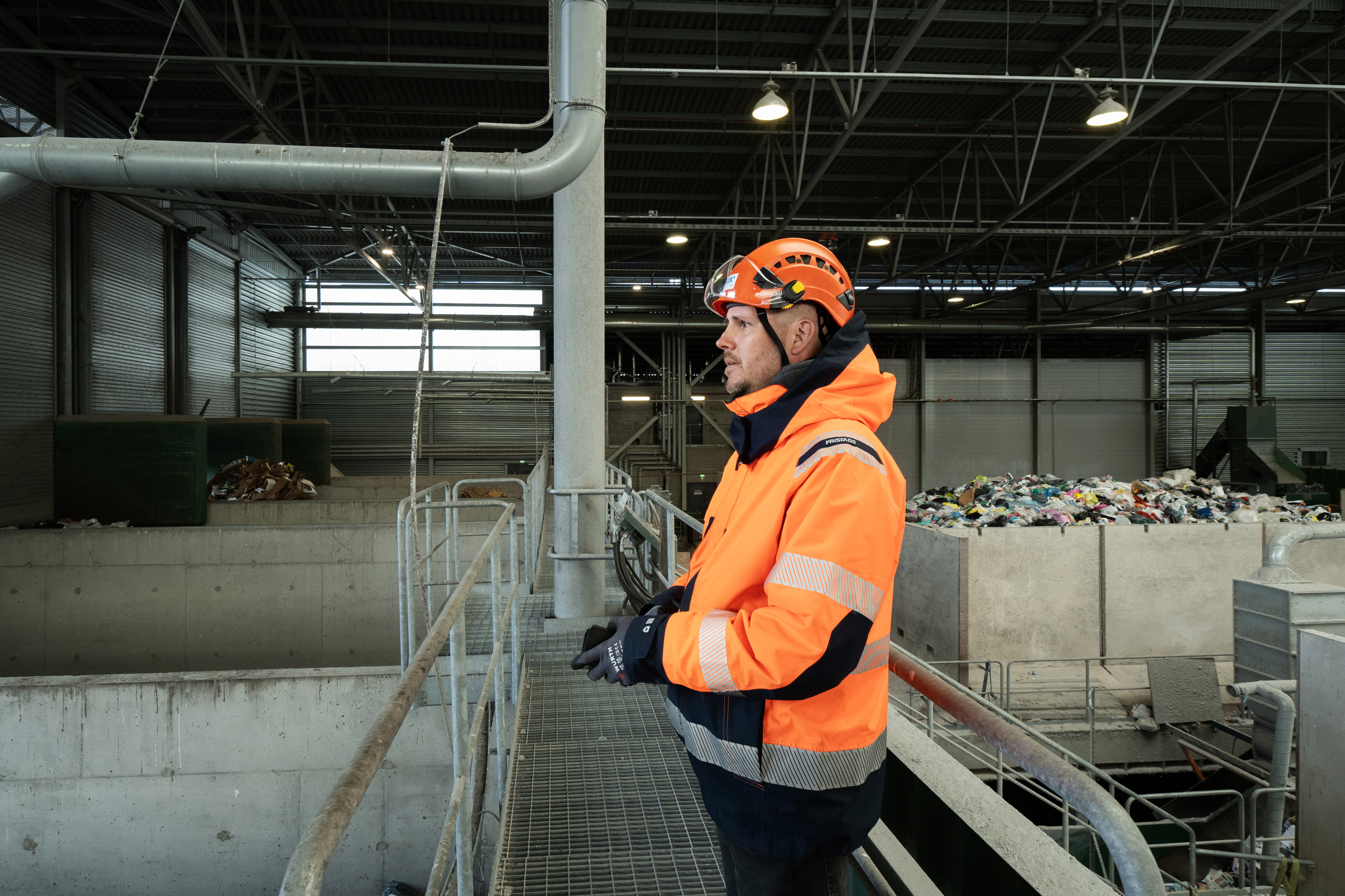 A male Stena Recycling employee in hi-vis protective gear, standing on the landing in a recycling facility looking to the side.