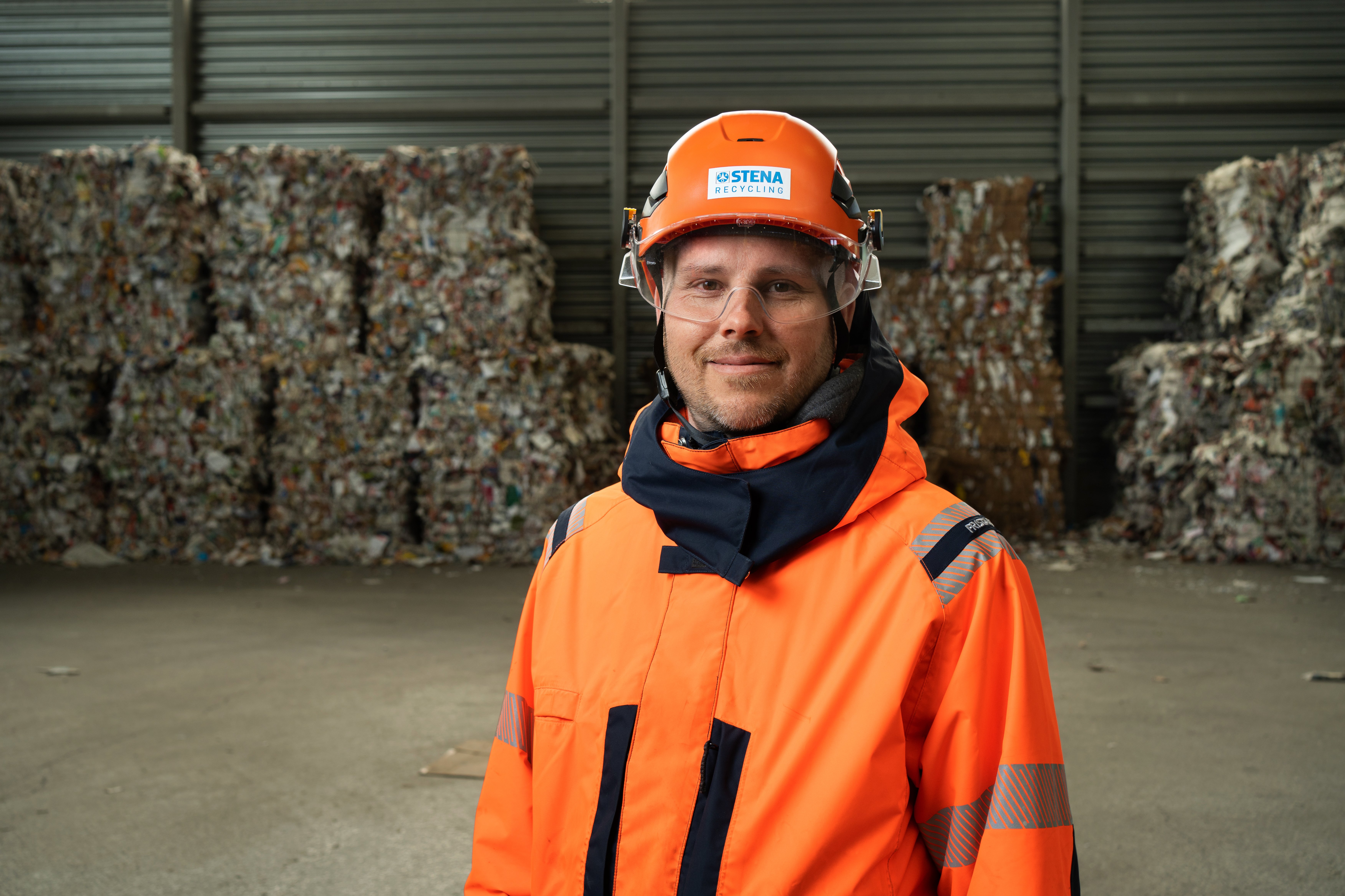 Close-up of a male Stena Recycling employee in hi-vis protective gear, standing in front of a piles with packaged paper waste